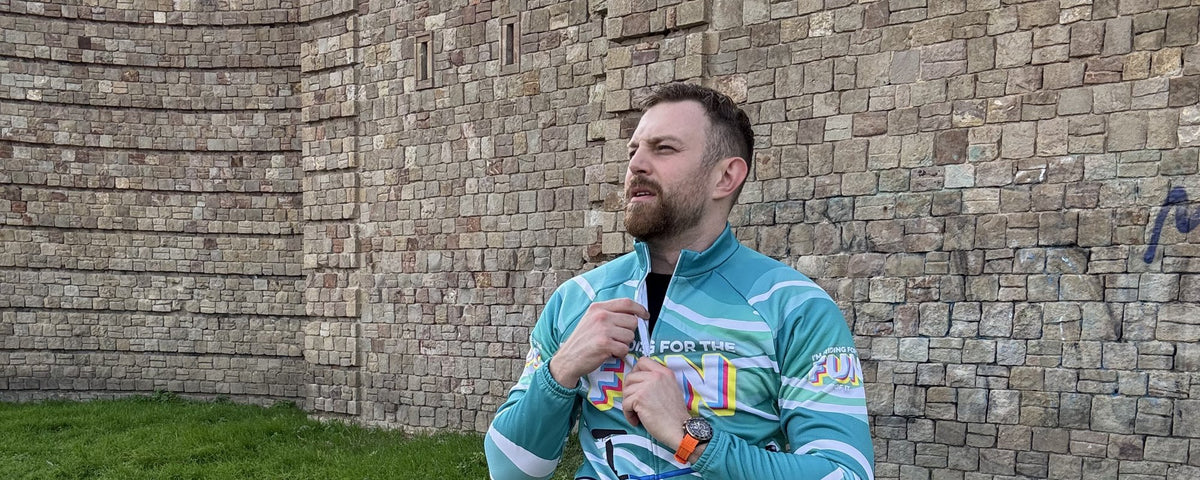 Rider in a teal long sleeve novelty cycling jersey and black bib shorts zipping up beside an old stone wall before a ride