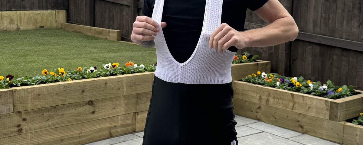 Rider holding the white mesh straps of black bib knicks over a black base layer, in a garden with flower beds