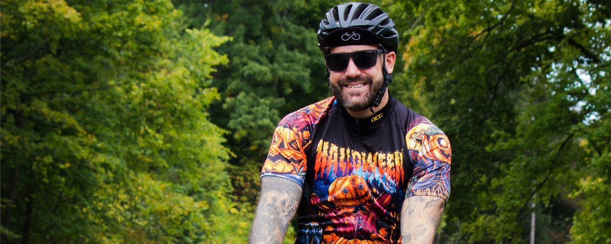 Smiling rider in a helmet and sunglasses on a tree-lined path, wearing a black jersey printed with Halloween lettering and jack-o'-lanterns