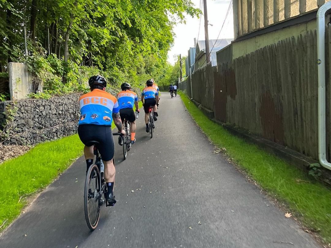 A line of riders in matching blue and orange jerseys on a tree-lined bike path, seen from the back of the group