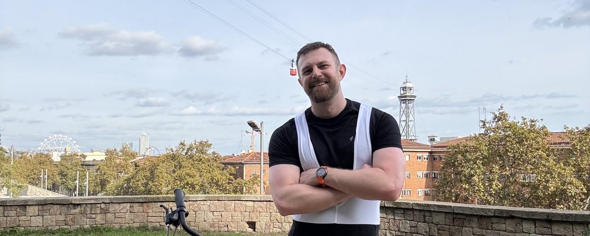 Smiling rider with arms crossed beside a hybrid bike in a park, wearing black bib shorts with white mesh straps over a black t-shirt