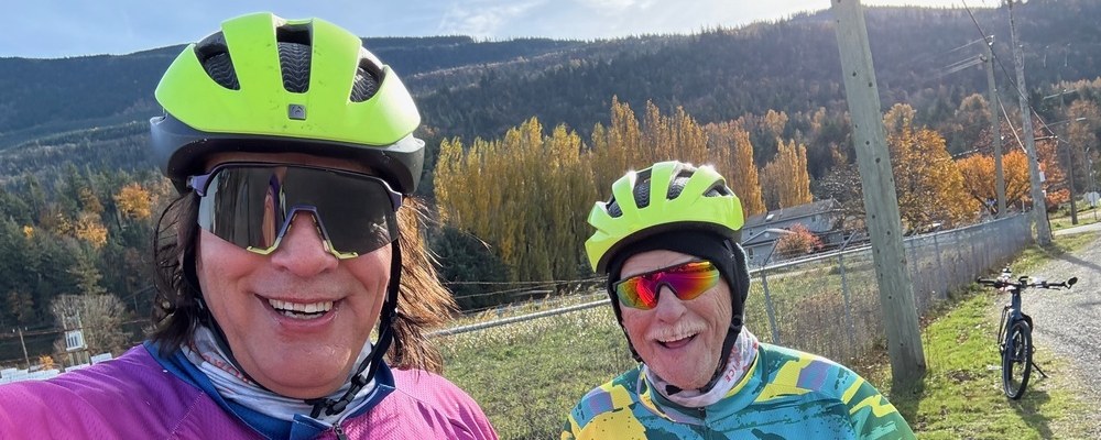Two smiling riders in helmets, sunglasses and long sleeve jerseys on an autumn roadside, one in a pink Toronto jersey and one in a yellow Yeah, Nah jersey