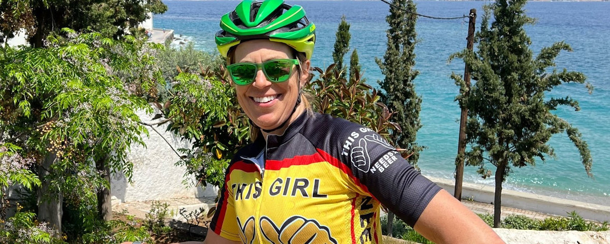 Smiling rider in a yellow and black This Girl Needs a Beer jersey leaning on a wall above the sea, bike beside her