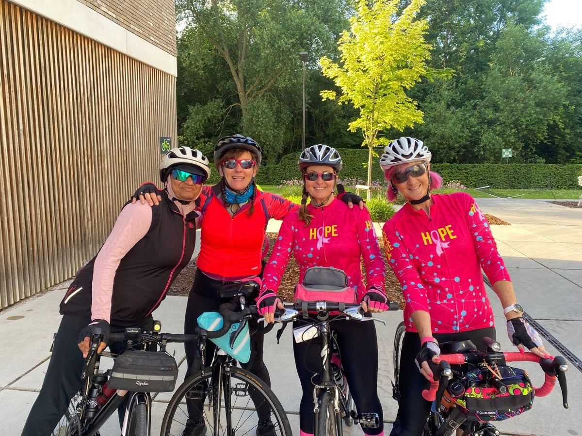 Four women riders with road bikes outside a building on a sunny morning, two of them in hot pink long sleeve jerseys printed with the word Hope and a pink ribbon