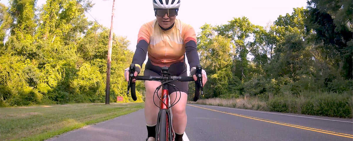 Rider in a white helmet and sunglasses riding a red road bike straight at the camera on a quiet tree-lined road, wearing an orange and peach sunset print women's cycling jersey with dark sleeves and black cycling shorts