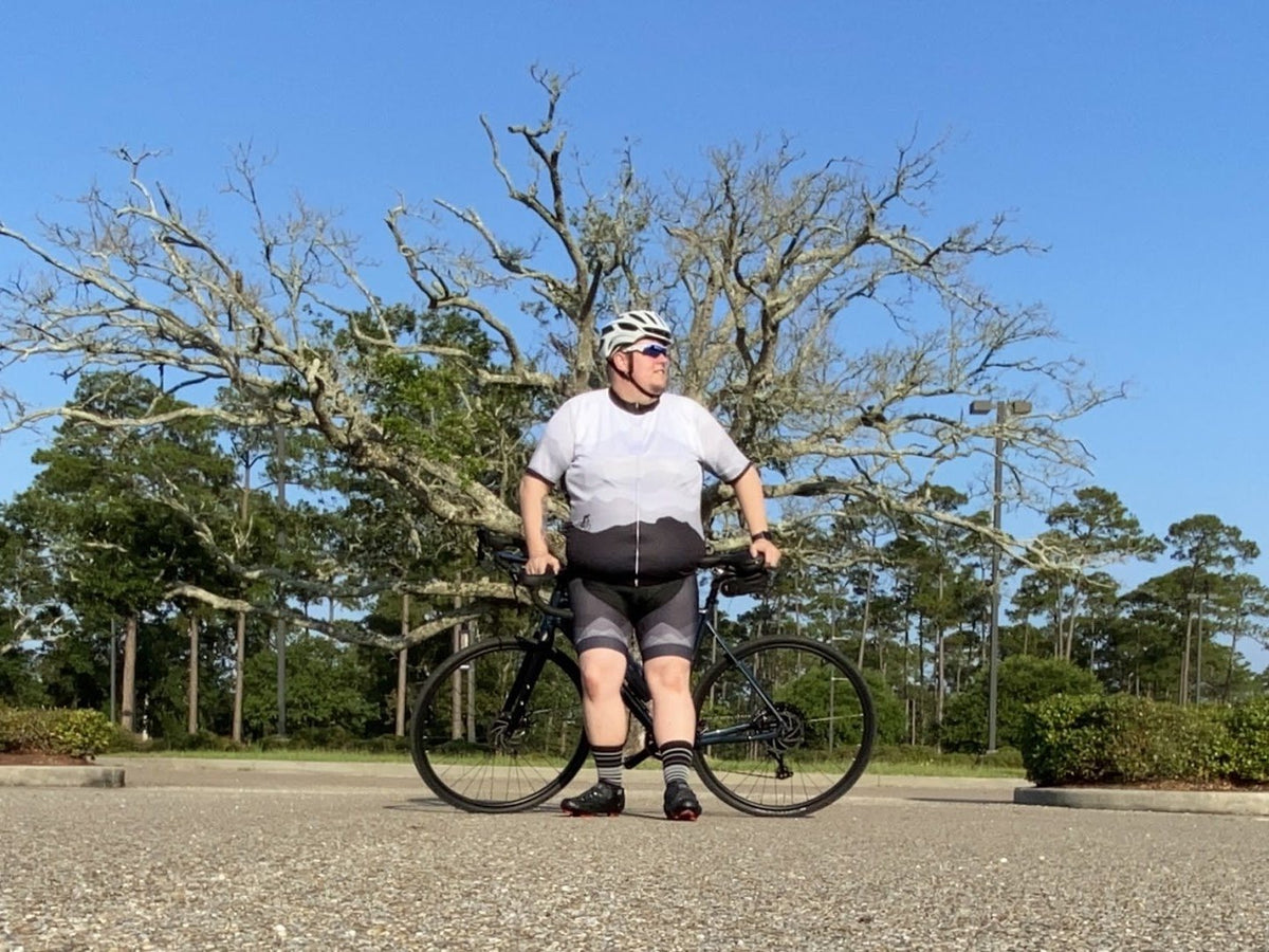 Rider in a white and grey jersey and matching padded cycling knicks, standing astride a road bike in a car park with a large bare tree and blue sky behind