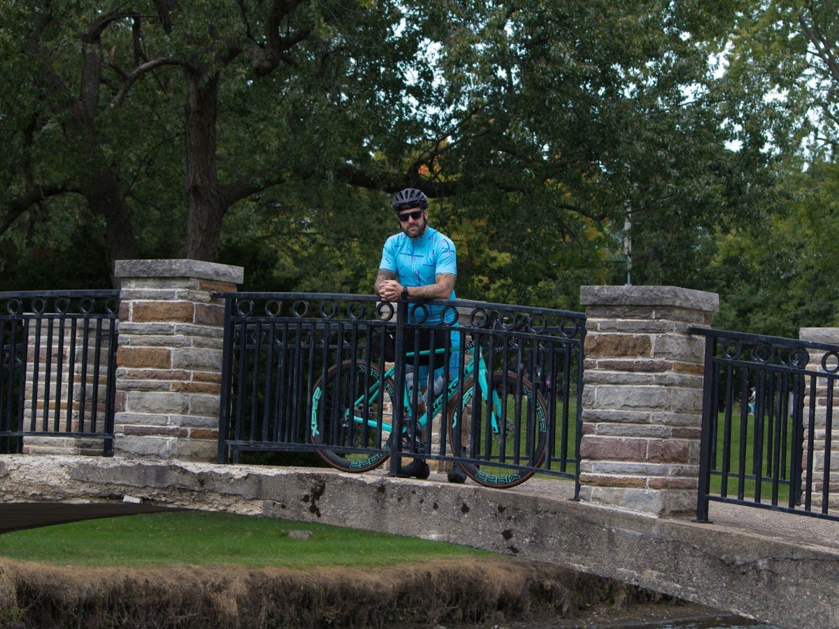 Bearded rider in a light blue jersey and black helmet leaning on the railing of a stone footbridge in a leafy park, with his bike beside him