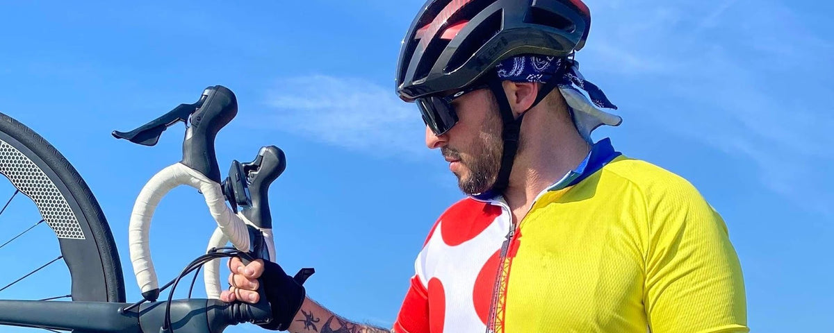 Rider on a beach holding a road bike up by the handlebars, wearing a jersey with yellow, green, white and red polka dot panels inspired by the Tour de France leader's jerseys