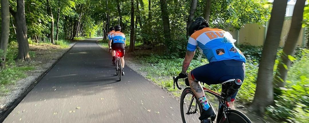 Two riders on a tree-lined bike path seen from behind, jersey back pockets visible
