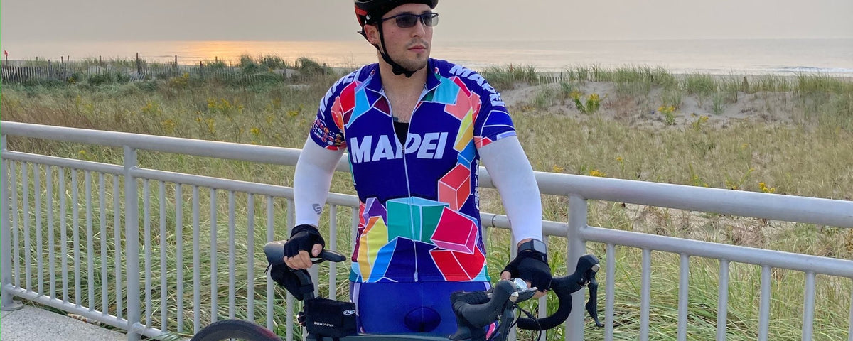 Rider in a multicoloured retro Mapei style long sleeve jersey standing with a road bike by a seaside railing