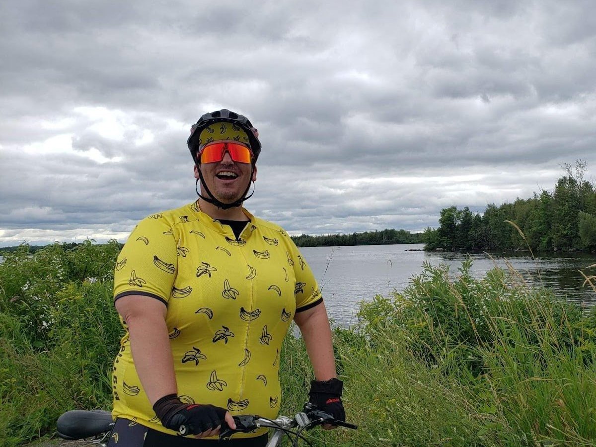 Laughing rider in a yellow banana print cycling jersey, orange mirrored sunglasses and a helmet, standing with a bike on a grassy path beside a lake under a cloudy sky