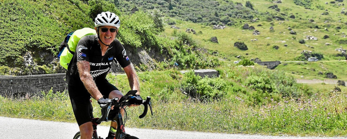 Smiling rider in a black jersey and black knicks climbing a sunny mountain road, arms shining with sweat, green hillside behind